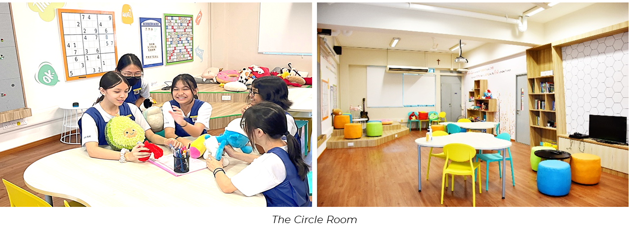 Students in uniforms sit around a table with stuffed toys beside a bright room with colorful chairs and shelves.
